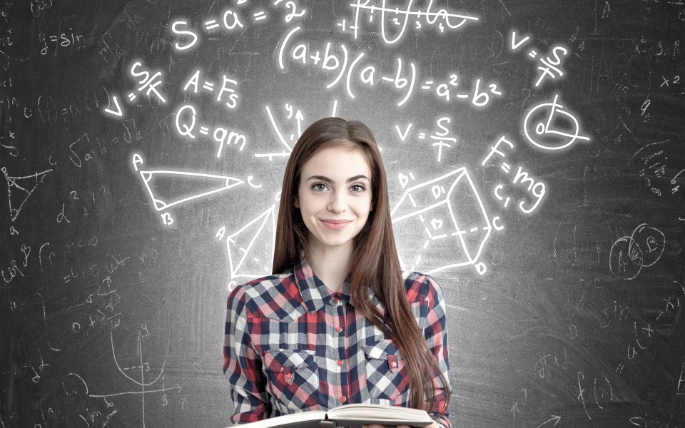 Portrait of a young girl holding an open book while standing near formulas written on a blackboard behind her.