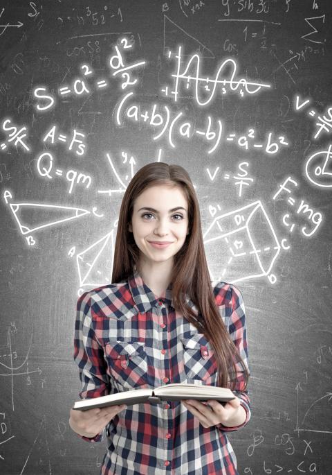 Portrait of a young girl holding an open book while standing near formulas written on a blackboard behind her.