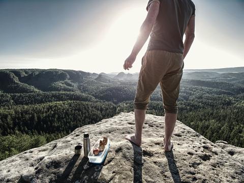 Man with his lunch and no shoes at top of cliff overlooking forest below
