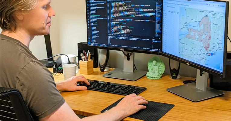 Nick at his desk with screens showing mapping and coding
